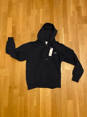 Helt ny Lacoste Zip-Hoodie - Säljer en klassisk Lacoste marinblå huvtröja med dragkedja i  nyskick. Plagget är aldrig använt och butikslappen sitter fortfarande kvar. Priset kan diskuteras.