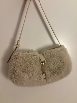 Beige fluffig axelremsväska - Supersöt beige handväska med fluffigt, mjukt material och justerbar axelrem. Väskan har guldfärgade metalldetaljer och ett spänne framtill som ger en trendig look. Perfekt accessoar för att lyfta din outfit med en cozy vibe.