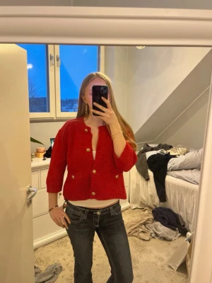 Röd kofta med guldknappar - Säljer en snygg röd kofta med guldknappar och trekvartsärmar. Koftan har ribbade detaljer vid ärmslut och nederkant samt två små fickor framtill. Perfekt att slänga över en topp för en cool look.