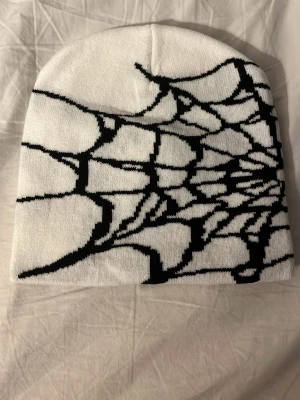 Vit mössa med svart spindelnätsmönster - Säljer en vit mössa med ett fett svart spindelnätsmönster runt hela. Mössan är mjuk och stretchig, perfekt för dig som vill sticka ut lite extra. Materialet känns som bomull eller akryl och designen är riktigt unik.