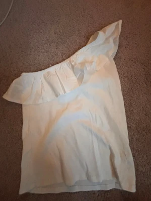 Vit oneshoulder topp med volang - Snygg vit oneshoulder topp med bred volang längs axeln. Perfekt för dig som vill ha en trendig och stilren look. Toppen är ärmlös och har en luftig passform som passar till många olika outfits.