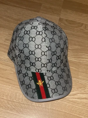 Grå keps med mönster och rand - Snygg grå keps med svart mönster av sammankopplade cirklar, samt en grön och röd rand med gul broderad insekt på skärmen. Justerbar rem baktill och böjd skärm. Materialet ser ut att vara canvasliknande tyg.