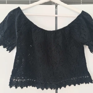 Svart offshoulder spetsblus - Trendig svart offshoulder blus i spets med korta ärmar och vågig kant nertill. Perfekt för dig som gillar en luftig och romantisk stil. Blusen har en croppad passform och är tillverkad i ett spetsliknande material. S/M