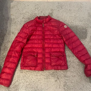 Röd dunjacka från Moncler - Snygg röd dunjacka från Moncler med klassisk quiltad design och två stora fickor framtill. Jackan har dragkedja hela vägen upp och Moncler-logga på ärmen. Perfekt för kyliga dagar när du vill hålla stilen.