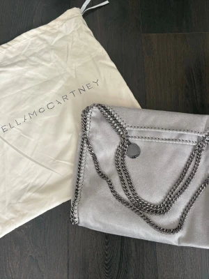 Grå väska - Snygg grå handväska liknande Stella McCartney Falabella. Väskan har kedjedetaljer i silver runt kanterna och som axelrem, samt en rund logotypbricka. ser nästan identisk ut med märken och ingraveringar, väldigt rymlig man får plats med dator