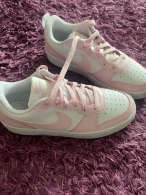 Nike vita och rosa låga sneakers - Säljer ett par fräscha Nike sneakers i vitt och ljusrosa. Skorna har klassisk låg siluett, perforerad tåbox och snörning. Tillverkade i syntetmaterial och textil med ikoniska swoosh-loggan på sidan. Perfekta för dig som vill ha en clean och trendig look.  Nypris 750