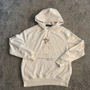 Vit hoodie Polo Bear från Ralph Lauren - Vit hoodie från Polo Ralph Lauren med ikoniskt Polo Bear-tryck på bröstet. Tröjan har huva med snörning och ribbade muddar. Materialet är mjuk bomull, perfekt för chill dagar. Klassisk och stilren design med coolt motiv framtill. Lite sliten på märket men inge mer än de.