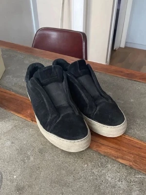 Svarta slip-on sneakers - Svarta slip-on sneakers med tjock vit sula och minimalistisk design. Ovandelen är i mockaliknande material utan synliga snören, vilket ger en clean och stilren look. Perfekta för dig som gillar enkelhet och streetstyle.