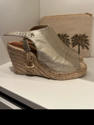 Guldiga kilklackade espadrillesandaler - Säljer ett par guldiga sandaler med kilklack och öppen tå. Ovandelen är i perforerat metallicläder och har justerbart spänne i guld. Klacken är i klassisk flätad jute, vilket ger en snygg espadrille-look. Perfekta för sommaren och ger extra höjd utan att tumma på komforten.