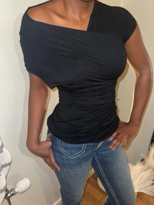 Svart oneshoulder topp - Snygg svart topp med oneshoulder-design och tight passform. Tyget är stretchigt och smickrande med draperade detaljer som ger en cool look. Perfekt för dig som vill sticka ut lite extra med en asymmetrisk stil.