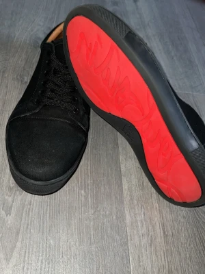 Svarta sneakers med röd sula - Snygga svarta sneakers från Christian Louboutin med klassisk röd sula och svarta skosnören. Ovandelen är i mocka och textil, och insidan har beige läderdetaljer. Perfekta för dig som vill sticka ut med exklusiv stil.