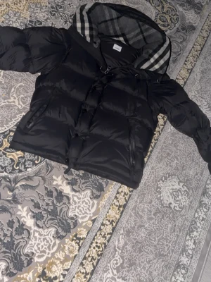 Den kända burberry jackan - Säljer en svart pufferjacka med dragkedja och knappar framtill. Stor huva med grått/vitt rutigt foder och två sidofickor med dragkedja. Varm och skön för vintern – och armarna går att ta av så den kan bli en väst.