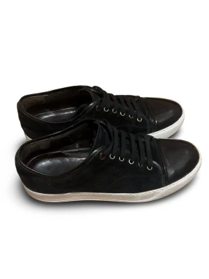 Svarta Lanvin Cap Toe  - Snygga svarta sneakers från Lanvin med klassisk design och vit sula. Skorna har svarta snören och metallfärgade öljetter. Materialet är mocha och har en glansig tå, vilket ger en stilren look. Perfekt för dig som gillar enkel och tidlös stil. Storlek 43, Priset är inte hugget i sten.