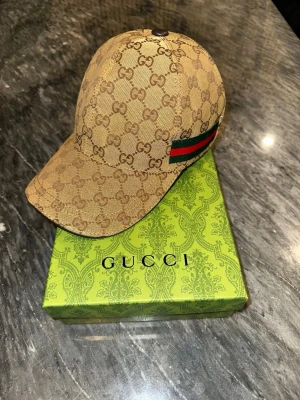 Beige keps från Gucci  - Säljer en beige keps från Gucci. På sidan finns en grön och röd rand som signaturdetalj. Kepsen är tillverkad i canvasmaterial och har böjd skärm. Perfekt för dig som gillar exklusiva accessoarer. Box och påse kommer med!