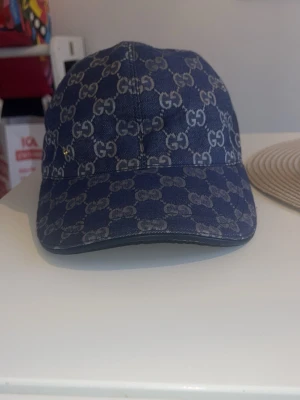 Blå denimkeps från Gucci - Snygg blå keps från Gucci i denim med det klassiska GG-mönstret över hela kepsen. Justerbar rem baktill i läder och böjd skärm. Perfekt för dig som vill ha en trendig accessoar med lyxig känsla.