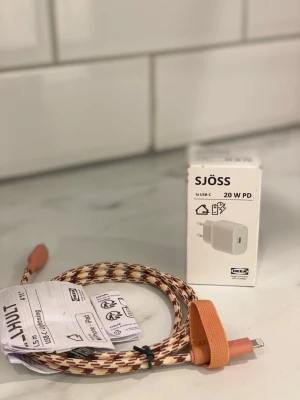 IKEA SJÖSS laddare & HULT kabel - Säljer en IKEA SJÖSS 20W USB-C PD väggladdare tillsammans med en IKEA HULT USB-C till Lightning-kabel. Kabeln är flätad i beige, brunt och orange med orangea kontakter och kardborreband. Perfekt för att ladda din iPhone eller annan kompatibel enhet.