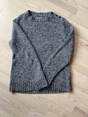 Gråmelerad stickad tröja med knappar - Säljer en gråmelerad stickad tröja med rund hals och tre svarta knappar på ena axeln. Tröjan har långa ärmar och ribbstickade muddar vid ärmslut och nederkant. Perfekt för kyliga dagar och enkel att matcha med jeans.