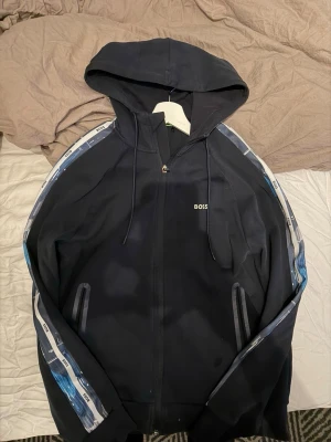 Svart hoodie från BOSS med blå detaljer - Svart hoodie från BOSS med dragkedja och huva. Ärmarna har blå och vita detaljer med BOSS-logga längs sidorna. Två fickor framtill och stilren design, perfekt till de flesta outfitsen. Priset ej hugget i sten så kom med förslag😊.
