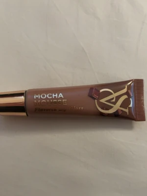 Mocha Mousse läppglans med chokladton - Säljer ett lyxigt läppglans från Victorias secret i färgen Mocha Mousse. Glanset har en mjuk brun nyans med chokladton och kommer i en elegant tub med gulddetaljer. Smaksatt och ger en glansig finish till läpparna. Perfekt för dig som gillar neutrala färger.