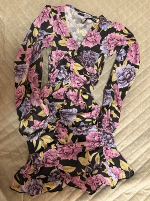 Blommig kortklänning med volang - Säljer en snygg kortklänning med blommigt mönster i lila, rosa, pastelgrön och svart på botten. Klänningen har v-ringning, volangdetaljer och långa ärmar. Perfekt för dig som gillar färgstarka plagg och vill sticka ut speciellt ny till våren eller påsken