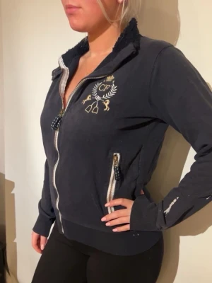 Mörkblå hoodie med dragkedja - Mörkblå hoodie med dragkedja framtill och broderad logga på bröstet. Fodrad krage i mjukt material och två fickor med dragkedja. Snygga detaljer i vitt och guld. Perfekt för en avslappnad och sportig stil.