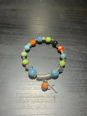 Färgglatt pärlarmband med korsberlock - Snyggt armband med pärlor i olika färger som blått, grönt, orange och svart. Armbandet har detaljer i silverfärgad metall och en hängande korsberlock. Perfekt accessoar för dig som gillar unika smycken med lite edge.