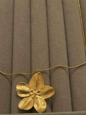Guldigt halsband med blomma - Snyggt halsband i guldfärg med en stor, detaljerad blomma som hänge. Kedjan är tunn och har ett justerbart lås med förlängningskedja. Perfekt för dig som gillar unika accessoarer med naturinspirerad design.
