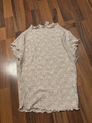 Beige spetsblus med volangkant - Säljer en beige spetsblus från H&M med korta ärmar och volangkant vid ärmslut och nederkant. Blusen har ett diskret blommigt mönster och är perfekt för dig som gillar romantiska detaljer och mjuka material. Storlek XS passar mer S!