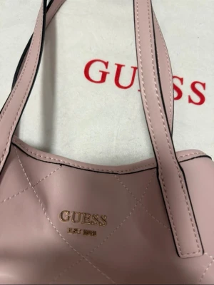 Rosa quiltad handväska från Guess - Säljer en ljusrosa handväska från Guess med quiltat mönster och guldfärgad logga framtill. Väskan har dubbla axelband och är tillverkad i slätt konstläder. Perfekt för dig som vill ha en stilren och trendig accessoar.