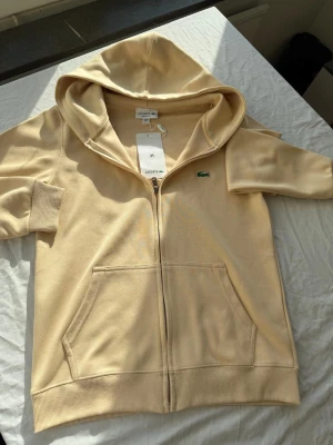 Beige hoodie med dragkedja från Lacoste - Snygg beige hoodie från Lacoste med dragkedja framtill och klassisk krokodil-logga på bröstet. Hoodien har stora fickor fram, ribbade muddar och en rejäl huva. Tillverkad i mjukt bomullsmaterial som känns skönt mot huden. Perfekt för en avslappnad och stilren look.