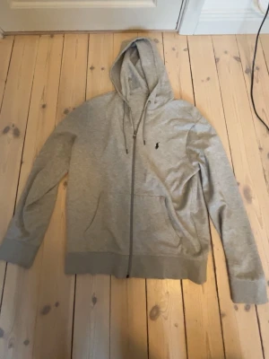 Grå zip hoodie från Ralph Lauren - Snygg grå hoodie med dragkedja från Ralph Lauren. Klassisk design med huva och dragsko, samt fickor framtill. Liten broderad logga på bröstet. Tillverkad i mjukt material som känns skönt mot huden. Perfekt för en avslappnad och stilren look.