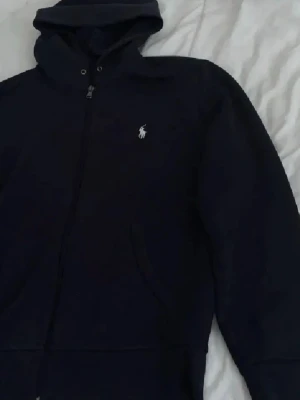 Mörkblå hoodie från Ralph Lauren - Snygg mörkblå hoodie från Ralph Lauren med dragkedja och klassisk vit logga på bröstet. Hoodien har huva med snörning och fickor framtill. Perfekt för en avslappnad och stilren look.