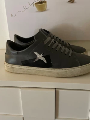 Svarta sneakers från Arigato bird sneaker grey - Snygga svarta sneakers från Axel Arigato i läder med vit sula och silverfärgad fågel på sidan. Klassisk låg modell med svarta skosnören och diskret logga. Perfekta för dig som gillar stilrena och trendiga skor.