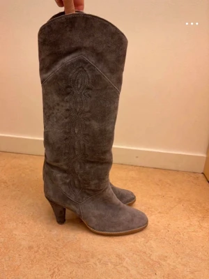 Grå mockastövlar Oswald - Snygga grå stövlar från Oswald i mocka med dekorativt mönster längs skaftet. Storlek 37. Ena klacken kan behöva klackas om men de funkar bra även utan, se bild. Därav priset.