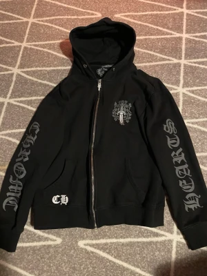 Svart Chrome Hearts hoodie med tryck - Säljer en svart hoodie från Chrome Hearts med dragkedja, huva och stora tryck på båda ärmarna. Bröstet har ett detaljerat Chrome Hearts-emblem och fickor framtill.