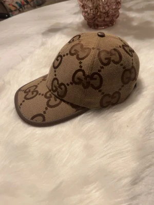 ÄKTA GUCCI JUMBO BEIGE - Snygg beige keps från Gucci med det klassiska Jumbo GG-monogrammet i brunt över hela kepsen. Justerbar rem baktill i läder och böjd skärm. Tillverkad i canvasmaterial som ger en exklusiv känsla och streetig look.