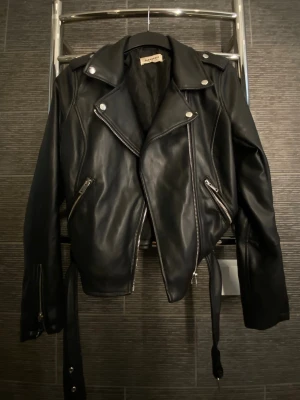Svart skinnjacka från NA-KD - Säljer svart skinnjacka från NA-KD med silverfärgade dragkedjor och knappar. Kommer även med ett bälte. 