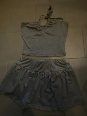 Grått set med topp och kjol - Säljer ett grått set i mjukt stretchigt material, består av en ärmlös topp med knytband i halsen och matchande kjol. 