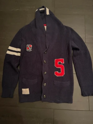 Mörkblå vintage wooden barell cardigan - Mörkblå stickad kofta från Wooden Barrel med stora knappar, två fickor fram och bred sjalkrage. Har vita ränder på ena ärmen, ett rött S-tryck och ett WB-emblem på bröstet. Perfekt för en sportig och avslappnad stil. Otroligt skön och bekväm, pris är diskuterbart som alltid!