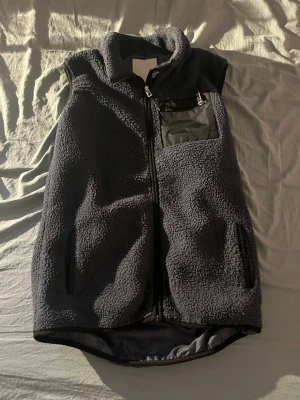 Mörkblå fleeceväst med dragkedja - Mysig mörkblå fleeceväst med hög krage och hel dragkedja framtill. Västen har två sidofickor med dragkedja och en bröstficka i tyg med dragkedja för extra förvaring. Perfekt för lager-på-lager och chill stil.