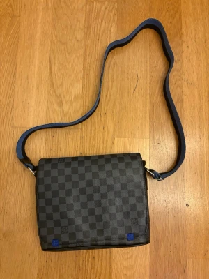 Louis Vuitton axelväska Damier Graphite - Snygg axelväska från Louis Vuitton i klassiskt Damier Graphite-mönster. Väskan är svart och grå med rutigt tryck och har en justerbar axelrem i blått och svart tyg. Perfekt storlek för det viktigaste och stängs med lock.