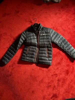 Svart dunjacka från BRL Racing - Snygg svart dunjacka från sail Racing med orange detaljer vid dragkedjor och insida. Jackan har hög krage, två sidofickor med dragkedja och quiltad design. Perfekt för kalla vinterdagar och ger en sportig look.