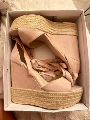 Platåsandaler med snörning - Säljer ett par trendiga platåsandaler med kilklack och snörning runt ankeln. beige/rose  färg. Sandalerna har en flätad sula i repstil och ovandel i mockaimitation. Perfekta för dig som vill ha extra höjd och en cool look. Aldrig använd 