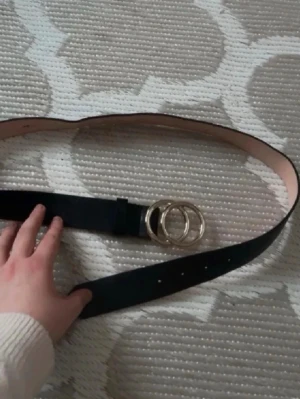 Svart bälte med dubbelring-spänne -  svart bälte med ett dubbelring-spänne i guld. Bältet är gjort i skinnmaterial. 