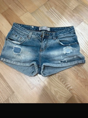 Blå lågmidjade jeansshorts - Snygga blå denimshorts med slitna detaljer och upprullade benslut. Klassisk femficksmodell med knapp och dragkedja framtill. Dom är lågmidjade🤩💕Kan tänka mig gå ner lite i pris 