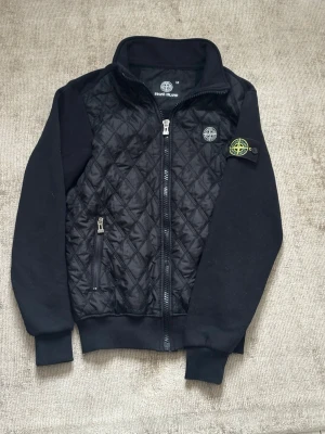 Svart cardigan från Stone Island - Svart cardigan från Stone Island med quiltad front och ärmar i mjuk bomull. Jackan har dragkedja framtill, ribbade muddar och den klassiska Stone Island-loggan på ärmen. Perfekt för dig som gillar snygga detaljer. Passar s och priset är ej hugget i sten