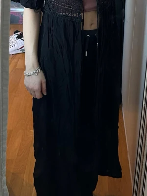 Svart strandklänning med spetsdetaljer - Svart strandklänning med v-ringning och vackra spetsdetaljer över bysten och midjan. Lätt och luftig modell med korta ärmar, perfekt att slänga över bikinin på stranden. Snygg och enkel att styla till sommarens alla stranddagar.