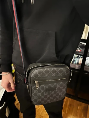 Gucci axelväska - Snygg axelväska från Gucci i mörkgrått med klassiskt GG-monogram över hela väskan. Väskan har en justerbar axelrem i rött och blått, en dragkedjeficka framtill och silverfärgade detaljer. Perfekt för att bära det viktigaste med stil.