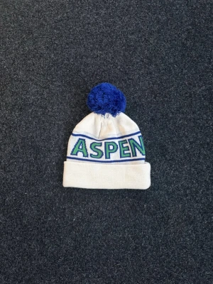 Vit mössa med blå tofs och ASPEN-tryck - Säljer en vit mössa med blå tofs och texten 'ASPEN' i blått och grönt. Mössan har vikt kant och är stickad i mjukt material, perfekt för kalla dagar. Snygg retrostil som sticker ut.