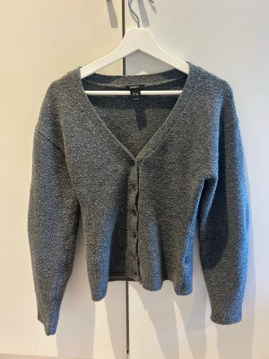 Grå kofta med v-ringning från Lindex - Mysig grå kofta från Lindex med v-ringning och knappar framtill. Koftan har lång ärm och är tillverkad i ett mjukt, lite fluffigt material som känns skönt mot huden. Perfekt att slänga över en topp för en avslappnad look.